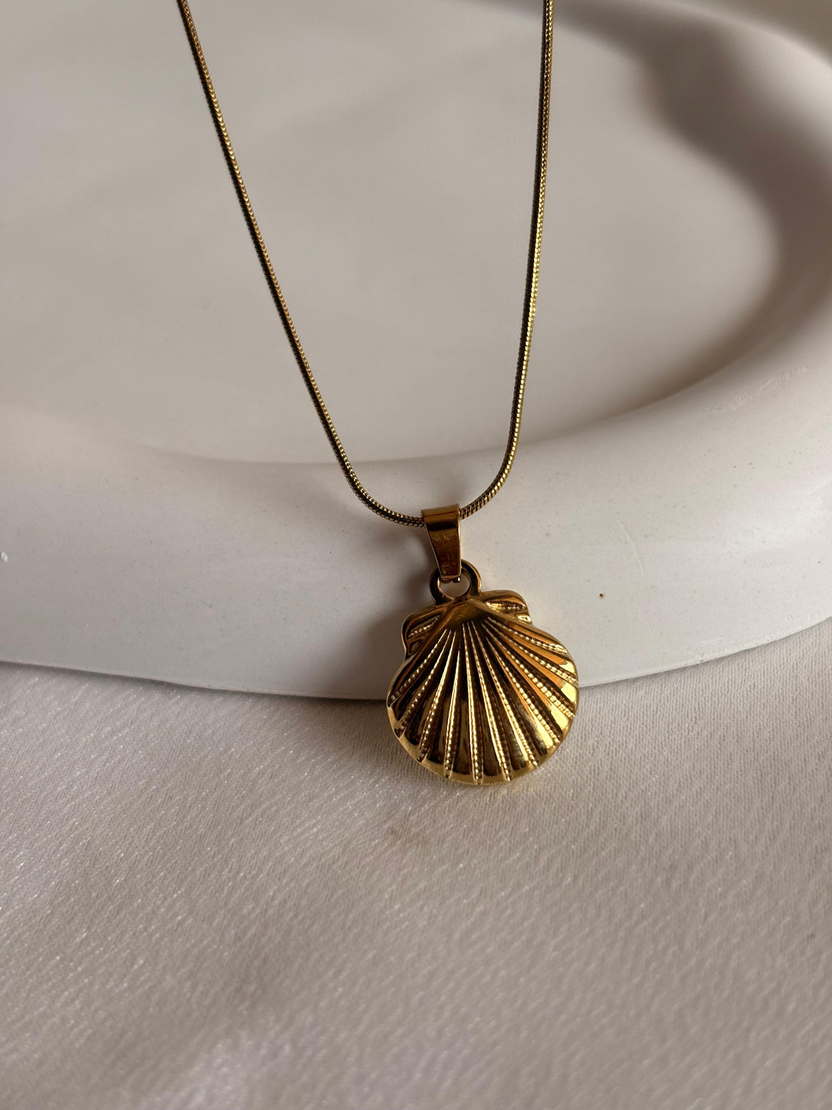 A gold shell-shaped pendant necklace resting on a white ceramic dish against a white fabric background.